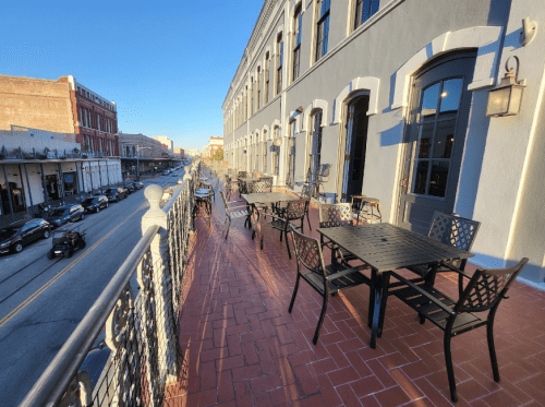 Outdoor dining area with black tables and chairs overlooking a street, bathed in warm sunlight.