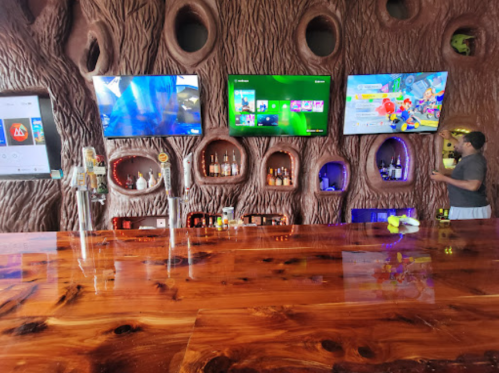 A wooden bar with multiple TVs displaying games, surrounded by unique, textured wall features and shelves of bottles.