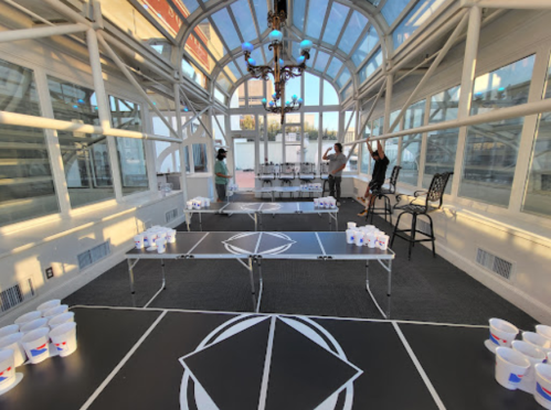 A bright indoor space set up for a beer pong game, with tables and cups arranged, and people preparing in the background.