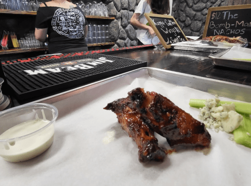 A plate with BBQ wings, celery, and dipping sauce, with a bar and menu in the background.