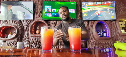 A bartender prepares colorful drinks at a bar with video game consoles and a TV in the background.