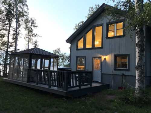 A cozy cabin with large windows, a deck, and a screened gazebo, surrounded by trees at dusk.