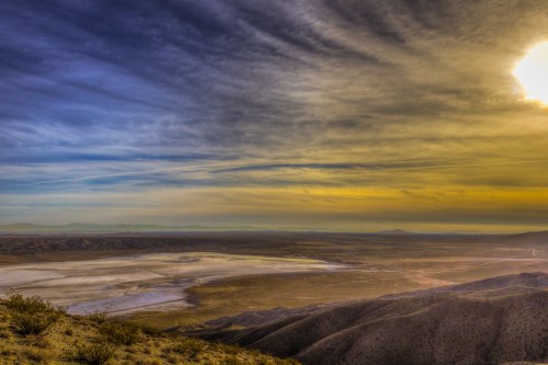 A vast desert landscape under a colorful sky, featuring mountains in the distance and a dry, cracked earth foreground.