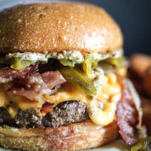 Close-up of a juicy burger with bacon, cheese, jalapeños, and a creamy sauce, served with crispy fries.