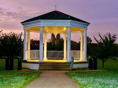 A lit gazebo with columns stands in a park at dusk, surrounded by trees and a pathway leading to it.