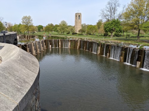 A serene park scene featuring a circular waterfall and a stone tower in the background, surrounded by greenery.