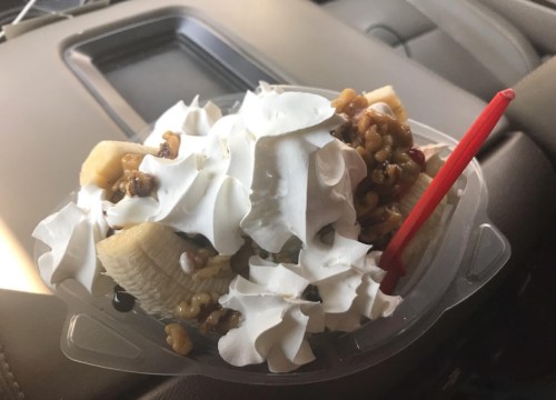 A dessert cup filled with whipped cream, banana slices, and nuts, sitting in a car.