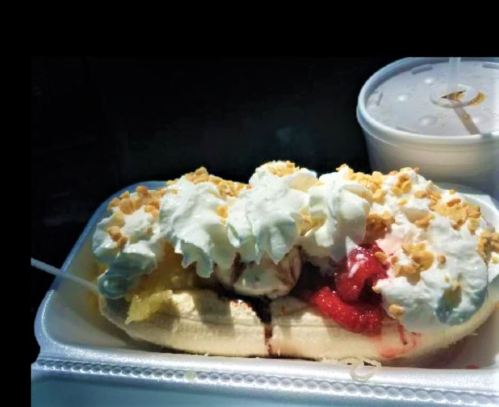 A banana split topped with whipped cream, nuts, and strawberry sauce, served in a takeout container.