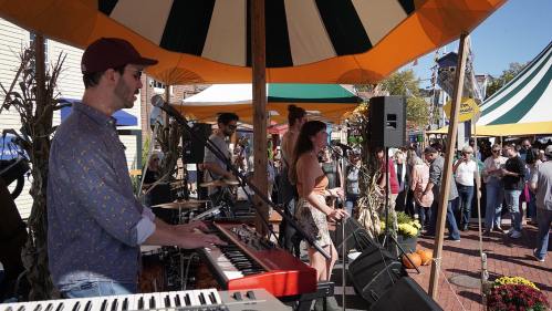A band performs under colorful tents at an outdoor festival, with a crowd enjoying the music in the background.