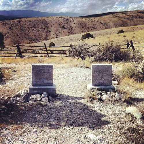 Two weathered gravestones stand on a rocky path, surrounded by dry grass and distant hills under a cloudy sky.