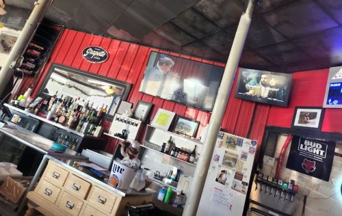 Interior of a bar with a red wall, shelves of liquor, and a TV showing a program. Decor includes framed photos.