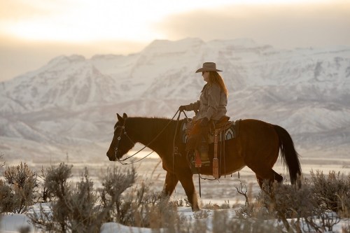 A woman rides a horse through a snowy landscape with mountains in the background during sunset.
