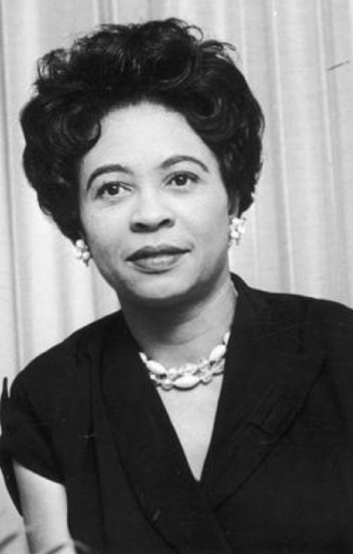 Black and white portrait of a woman with curly hair, wearing a dark outfit and pearl jewelry, looking directly at the camera.