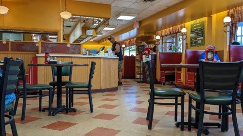 A diner interior with red booths, tables, and a counter; a few customers are seated and one is at the counter.