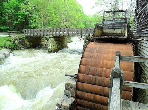 A wooden water wheel beside a rushing river, with a bridge in the background and lush greenery surrounding the scene.