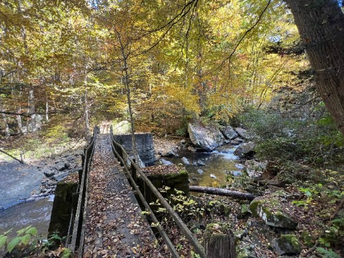 A wooden bridge crosses a stream surrounded by colorful autumn foliage and rocky terrain.