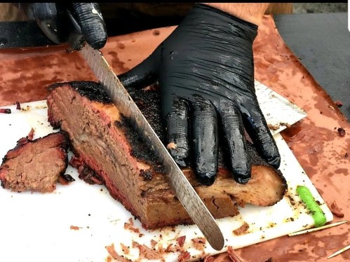 A person in black gloves slices a smoked brisket on a cutting board, with pieces of meat scattered around.