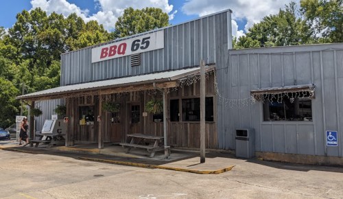 Exterior of BBQ 65 restaurant with wooden benches, a sign, and greenery under a blue sky with clouds.