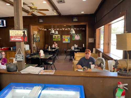 A cozy restaurant interior with wooden walls, tables, and patrons enjoying their meals. Decor includes artwork and a pig statue.
