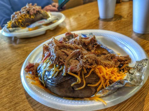 A loaded baked potato topped with pulled pork and shredded cheese, served on a paper plate.