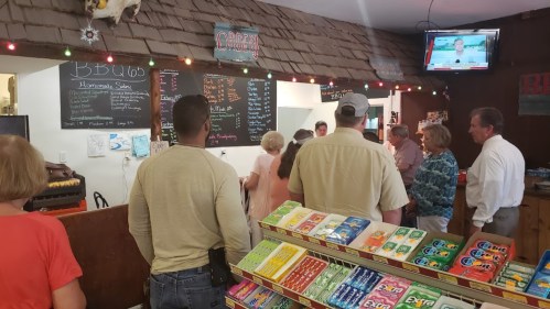 A busy BBQ restaurant with customers in line, colorful candy displays, and a TV showing news in the background.