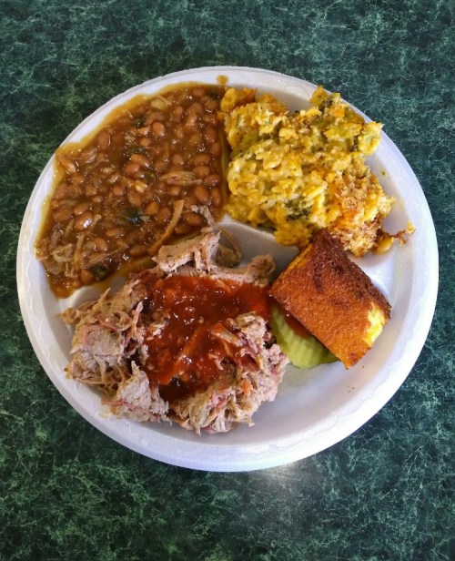 A plate with pulled pork topped with sauce, baked beans, cheesy broccoli rice, and cornbread on a green table.