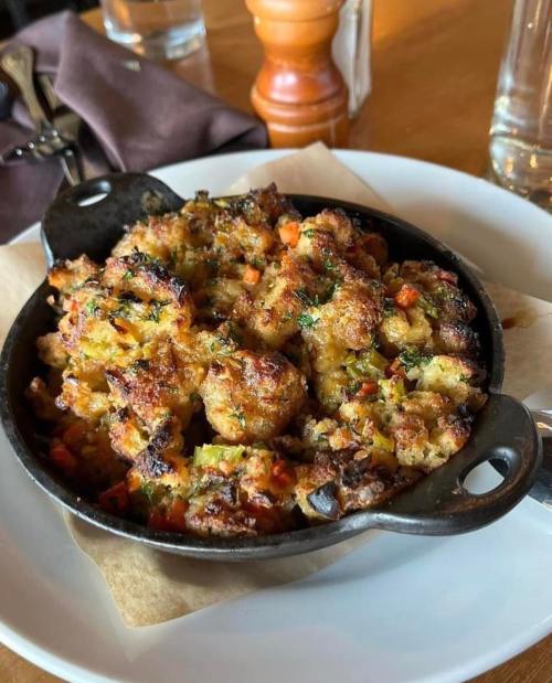 A cast-iron dish filled with golden-brown stuffing, featuring herbs and colorful vegetables, served on a white plate.