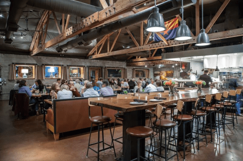 A bustling restaurant interior with wooden beams, a bar area, and diners enjoying meals at tables.