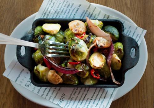 A cast-iron dish of roasted Brussels sprouts, red onions, and peppers, served on a white plate with newspaper underneath.