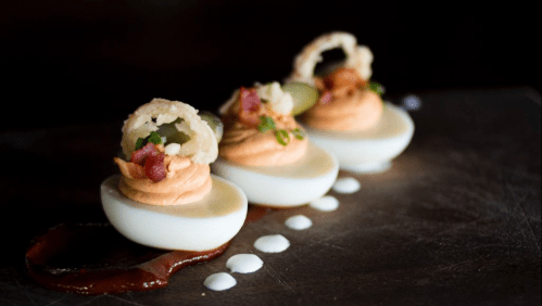 Three gourmet deviled eggs topped with crispy onion rings, bacon bits, and garnished with sauces on a dark plate.