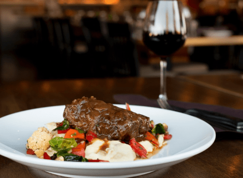 A plate of braised beef on a bed of mashed potatoes, surrounded by colorful vegetables, with a glass of red wine in the background.