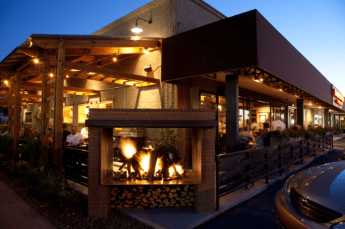 A cozy restaurant exterior at dusk, featuring outdoor seating, a fire pit, and warm lighting.