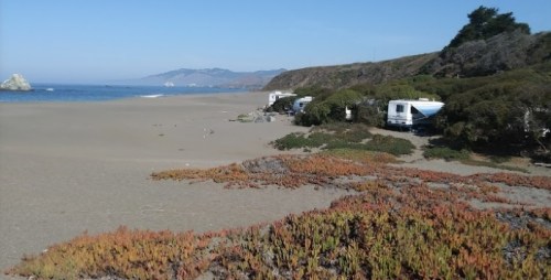 A sandy beach with RVs parked near the shore, surrounded by coastal vegetation and distant hills under a clear blue sky.