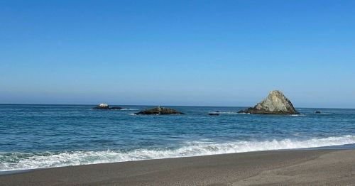 A serene beach scene with gentle waves, featuring two rocky outcrops in the calm blue ocean under a clear sky.