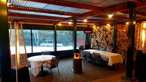 Cozy outdoor dining area with tables, heaters, and a stacked wood wall, surrounded by snow-covered scenery.