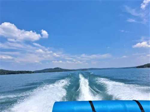 A view from a boat showing a wake on a calm lake under a blue sky with fluffy clouds and distant hills.