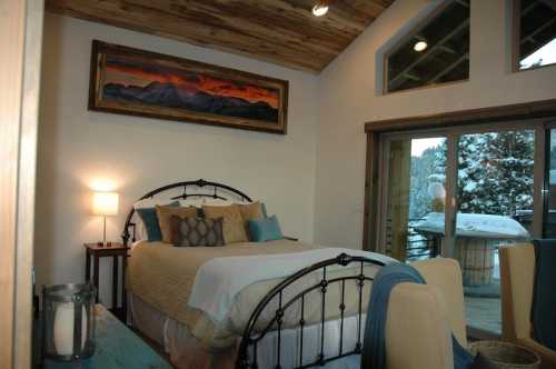 Cozy bedroom with a metal bed frame, warm lighting, and a mountain painting, featuring a snowy view outside.