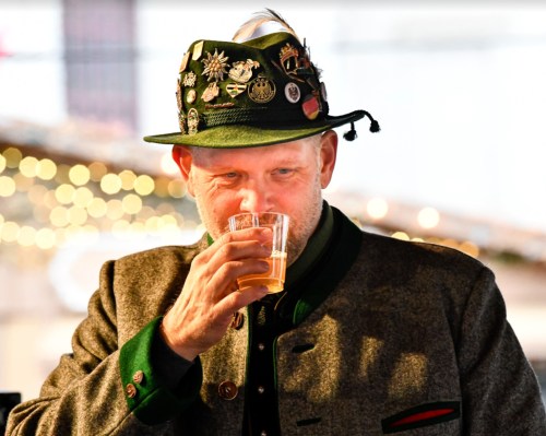 A man in a traditional hat and jacket sips a drink, surrounded by festive lights.