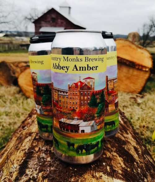 Three cans of Abbey Amber beer from Country Monks Brewing, featuring a colorful landscape design, set on a log outdoors.