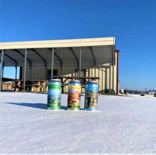 Three colorful cans of beer sit on a snowy surface in front of a metal building under a clear blue sky.