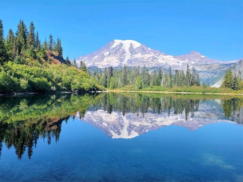 A serene lake reflects a snow-capped mountain and lush green trees under a clear blue sky.