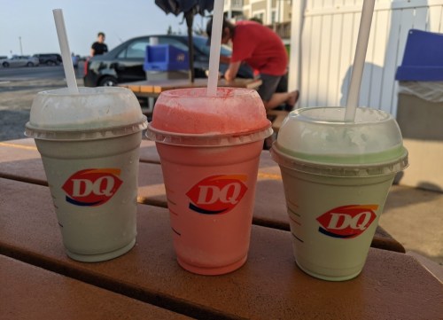 Three Dairy Queen cups with colorful milkshakes—green, pink, and light blue—sitting on a wooden table.