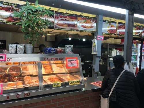 A food counter with a variety of meats displayed, and a customer waiting to order. Menu items are visible above.