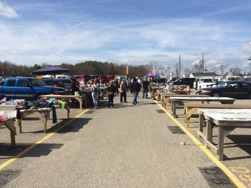 A bustling outdoor market with vendors' tables on either side and shoppers browsing under a clear blue sky.