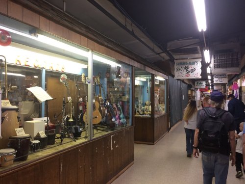 A bustling indoor market with music shops displaying guitars and instruments, and shoppers walking by.
