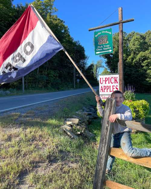 A child holds a large flag while sitting on a fence near a sign for a U-Pick apple orchard.