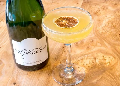 A sparkling cocktail garnished with a dried orange slice, next to a bottle of sparkling wine on a wooden surface.