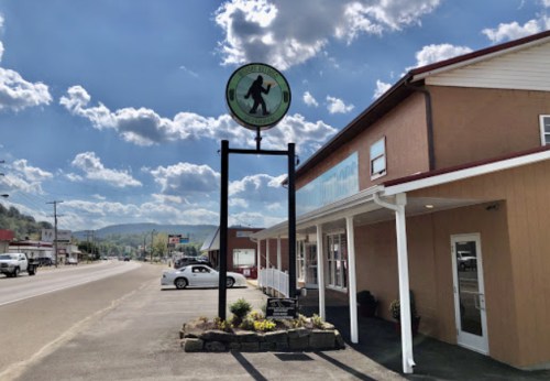 A sign featuring a Bigfoot silhouette stands outside a building on a sunny day, with a street and mountains in the background.