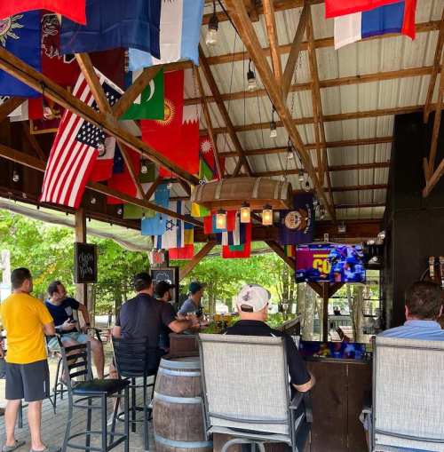 A lively bar with various flags hanging, patrons watching a game on TV, and a rustic wooden interior.