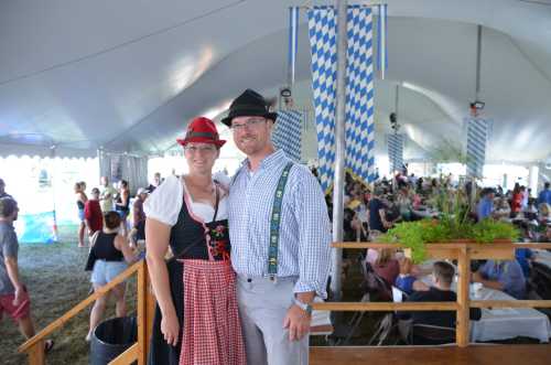 A couple in traditional attire poses inside a large tent filled with people enjoying a festive event.